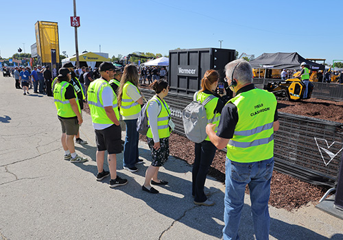 A group of people wearing bright yellow safety vests labeled 'Field Classroom' standing outdoors near a demonstration area with heavy equipment and machinery, surrounded by event tents and banners.