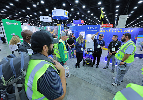A group of people in bright yellow safety vests gathered at a The Utility Expo show booth with banners and displays, listening to a demonstration near equipment and signage.