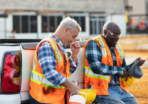 Two multiracial construction workers taking a coffee break, conversing, leaning on the tailgate of a pickup truck. The African-American man is in his 40s. The main focus is on his coworker, a senior man in his 60s.