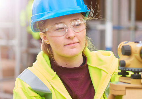 a female construction worker stands behind a builder's level on a building site .She is smiling proudly to camera.