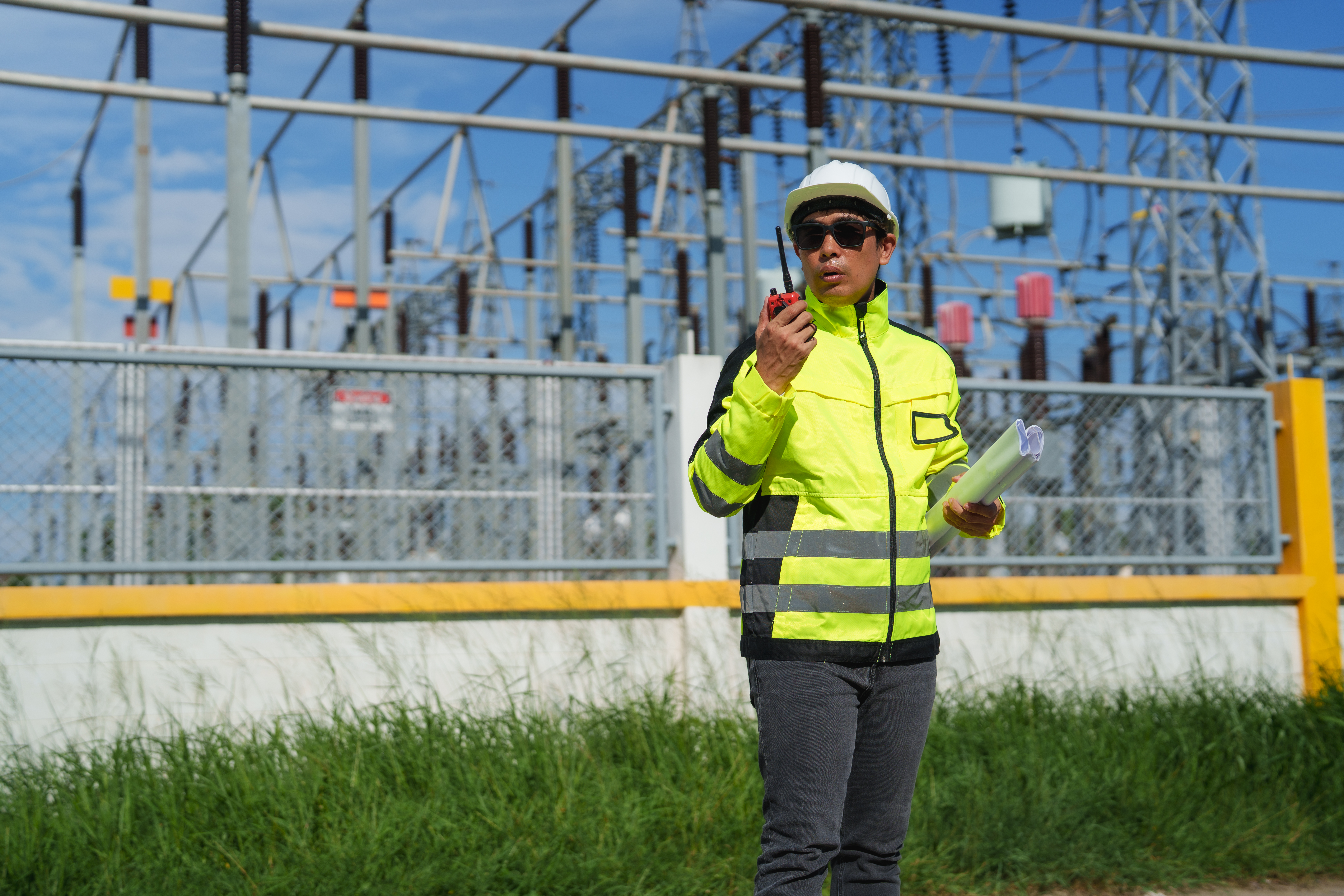 Electrical engineer standing at a power substation, holding blueprints and communicating via a walkie-talkie, wearing a hard hat and safety vest while overseeing critical infrastructure operations