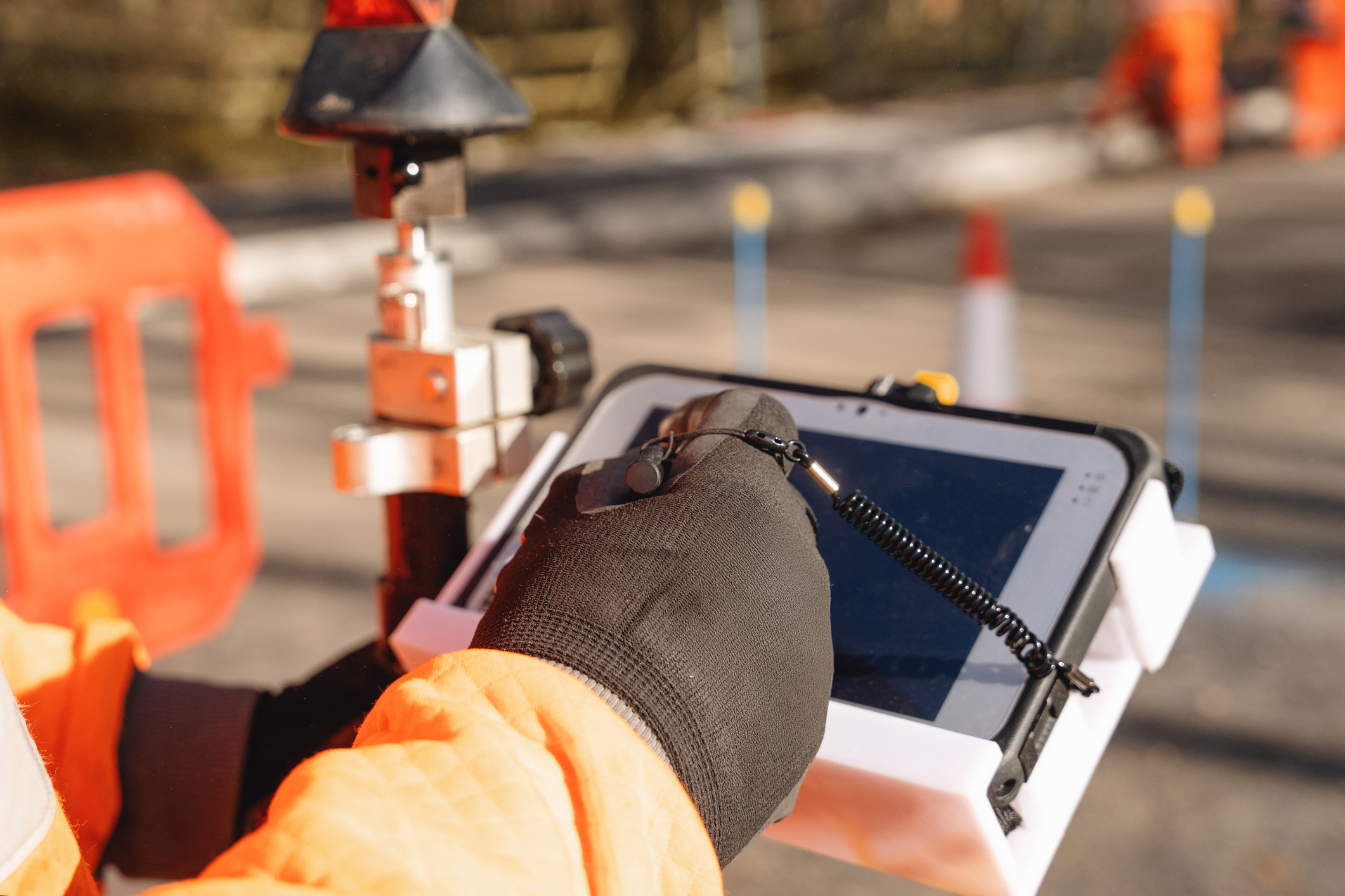 Close-up of construction worker in a bright orange uniform is focused on operating a tablet while standing near traffic cones and surveying equipment on construction site