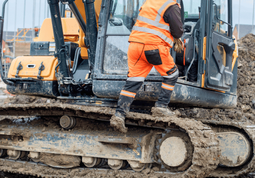 Construction worker in bright safety gear working hard using shovel to clean tracks of his heavy machinery excavator on muddy site