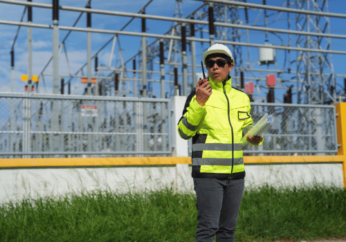 Electrical engineer standing at a power substation, holding blueprints and communicating via a walkie-talkie, wearing a hard hat and safety vest while overseeing critical infrastructure operations