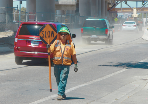 Milpitas, California, United States - July 29, 2016: The American worker in an orange vest shows sign "Slowly." Behind the cars, a lot of dust and bridge under construction.