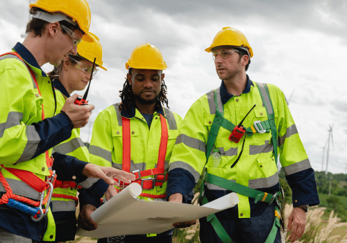 group of construction workers wearing yellow vests and hard hats are looking at a blueprint. They are discussing the details of the project and making sure everything is in order
