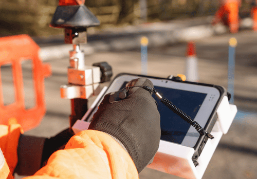 Close-up of construction worker in a bright orange uniform is focused on operating a tablet while standing near traffic cones and surveying equipment on construction site