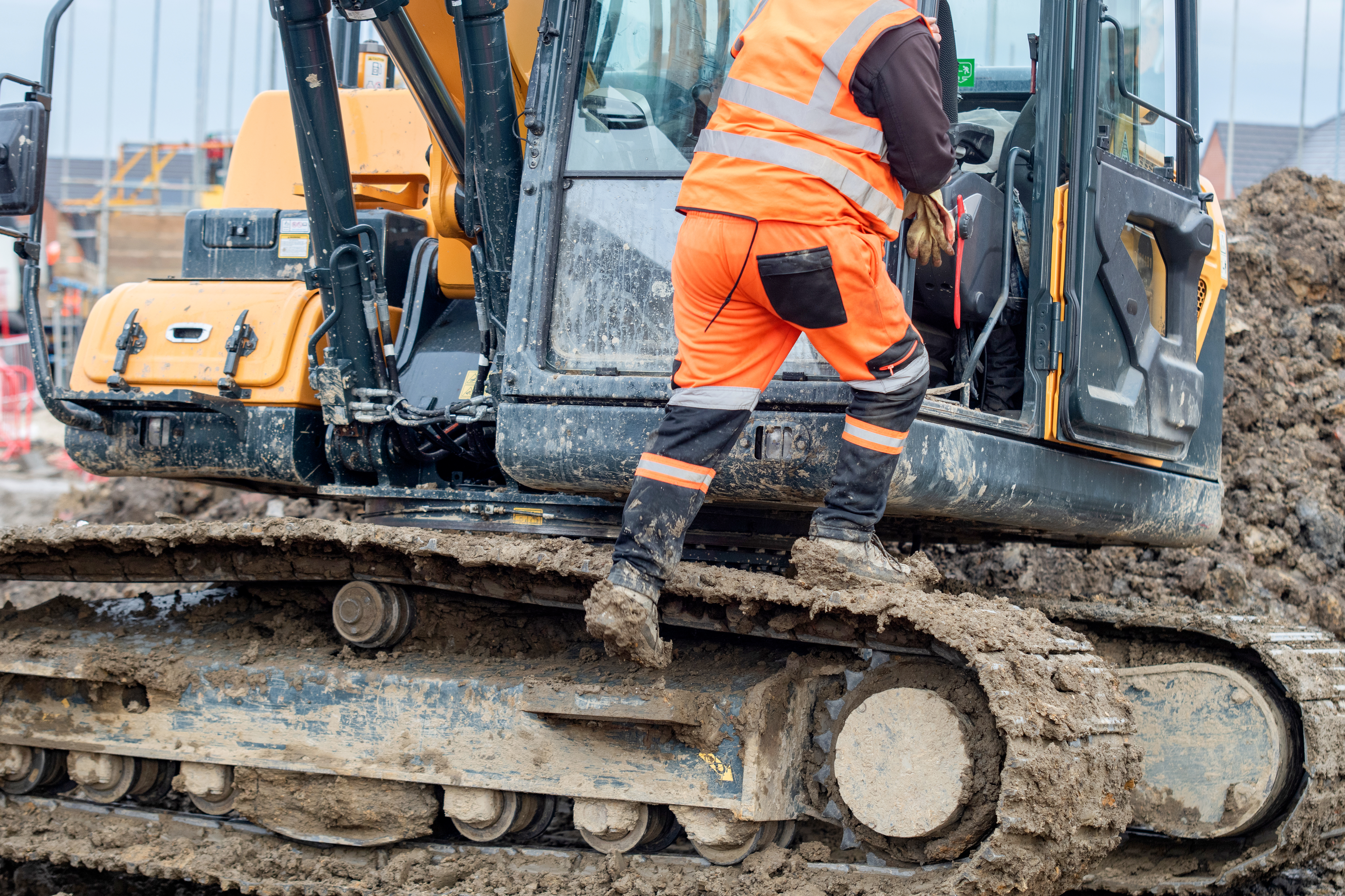 Construction worker in bright safety gear working hard using shovel to clean tracks of his heavy machinery excavator on muddy site