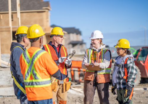 A small group of constructions workers meet with the forewoman to review the days work. They are each wearing proper safety equipment and are listening attentively.