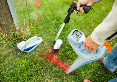 An electric utility worker is using a radio frequency detector to determine a buried power line location and mark it with spray paint and a red flag. The transmitter is to the left and is connected to the ground line from a utility pole. The can painter has some motion blur. This image represents all of the components as a result of, "call before you dig".