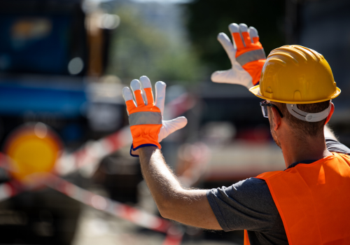 A construction worker in safety gear is directing vehicles at a construction site. Bright orange gloves and a helmet stand out against the background of equipment and barriers.