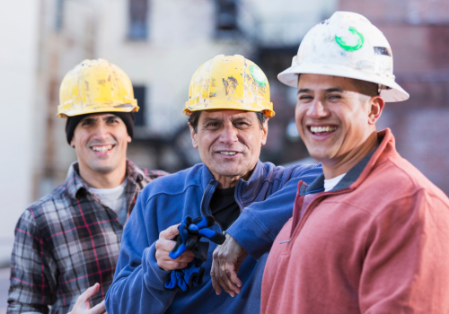 A group of three multi-ethnic men wearing white and yellow hard hats, standing in a row. The focus is on the worker in the middle, He is the foreman of this work crew, an Hispanic man in his 50s wearing a royal blue shirt, smiling and pointing at the camera. They are construction workers, engineers or utility workers in the city, buildings out of focus behind them.