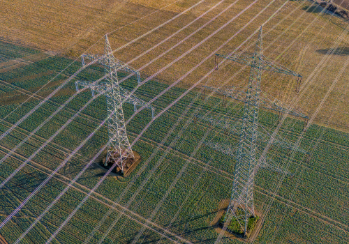Aerial drone view of high-voltage power transmission pylons stretching across a rural agricultural field. The image highlights energy infrastructure, electricity distribution, and the contrast between technology and nature in the countryside